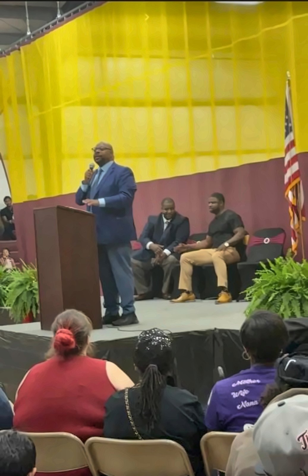 Kermit Williams speaking at a podium on stage with guests seated behind him